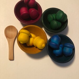 May include: A wooden toy set with four small, round bowls in red, green, yellow, and blue, each filled with acorn-shaped pieces. A small wooden spoon is placed beside the bowls. The toys are arranged on a white surface.
