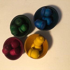 May include: Four small wooden bowls, each filled with acorn-shaped objects in different colours: green, blue, red, and yellow. The bowls are arranged on a white surface, creating a colourful and playful display.