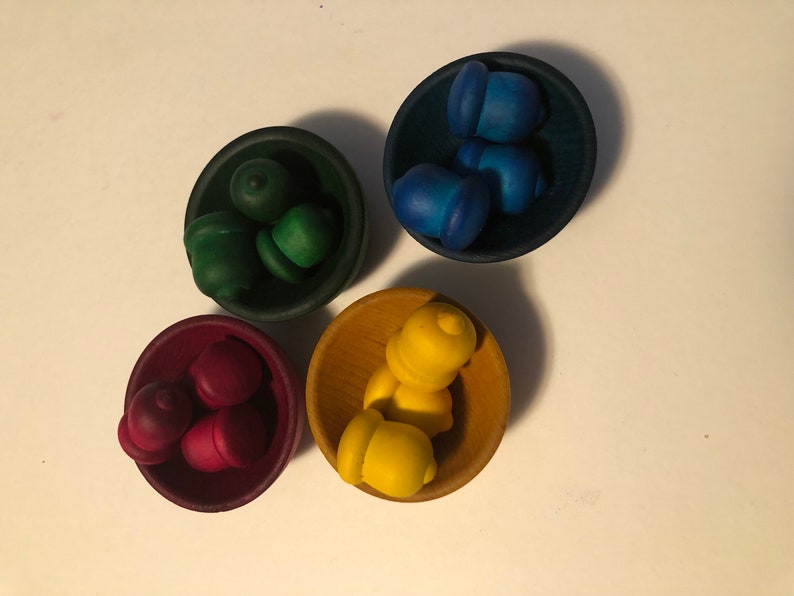 May include: Four small wooden bowls, each filled with acorn-shaped objects in different colours: green, blue, red, and yellow. The bowls are arranged on a white surface, creating a colourful and playful display.