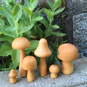 May include: Six wooden mushroom figurines of varying sizes, with natural wood tones, arranged on a gray concrete surface.