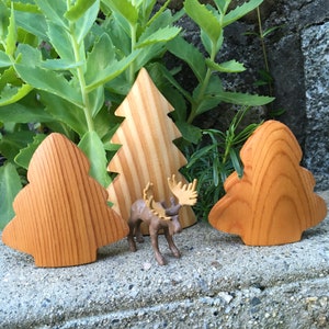 May include: Wooden Christmas tree decorations and a small moose figurine are arranged on a stone surface, with green foliage in the background. The trees are made of light-coloured wood with visible grain.