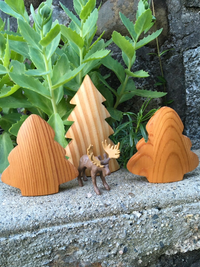 May include: Wooden Christmas tree decorations and a small moose figurine are arranged on a stone surface, with green foliage in the background. The trees are made of light-coloured wood with visible grain.
