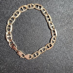 May include: A gold-coloured mariner chain bracelet. The bracelet features interlocking oval links, each with a bar across the centre. The clasp is a rectangular box style. The bracelet is displayed on a dark grey textured surface.