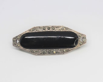 May include: A silver brooch featuring a black oval stone set within a geometric frame. The frame is adorned with small, sparkling stones.