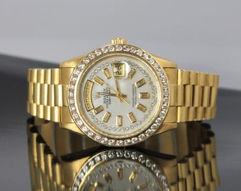 Puede incluir: Un reloj Rolex Oyster Perpetual Day-Date de oro con un bisel engastado con diamantes y una esfera de nácar blanco. El reloj tiene una ventana de fecha y una visualización del día de la semana.