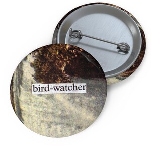 May include: A round button with a vintage-style image of a forest scene and the text "bird-watcher" printed on it.