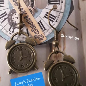 Può includere: Un paio di orecchini di ispirazione steampunk. Ogni orecchino presenta un piccolo ciondolo a forma di sveglia color bronzo con numeri romani sul quadrante. Gli orecchini sono sospesi a un gancio curvo. Il testo "Jane's Fashion & Art" è visibile.