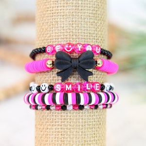 May include: A stack of four bracelets with pink and black beads. The top bracelet has the words "BE YOU" in white letters on pink beads. The second bracelet has a black bow. The third bracelet has the word "SMILE" in white letters on pink beads. The bottom bracelet has black and white beads.