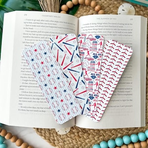 May include: An open book with several baseball-themed bookmarks. The bookmarks feature baseballs, bats, and stars in red, white, and blue patterns. The book is on a woven mat, with decorative beads and a tropical leaf in the background.