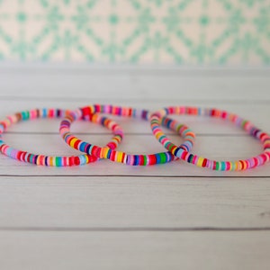 May include: Three colorful beaded bracelets stacked on a white wooden surface. The bracelets are made of small, round, multi-colored beads.