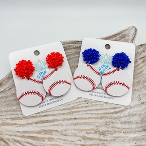 May include: A pair of baseball-themed earrings with red and blue beaded charms and white wooden baseballs. The earrings are displayed on a white card with the text "Follow us on IG and FB @EllieAndGingerCo".