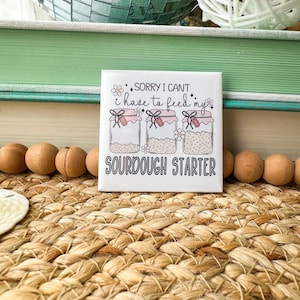 May include: A square ceramic tile with the text "Sorry I can't, I have to feed my sourdough starter." The tile features illustrations of three jars with pink lids and bows. The background includes a woven mat, wooden beads, and a decorative mirror.