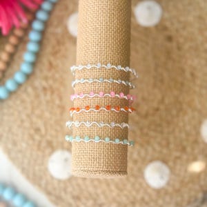 May include: A display of six beaded bracelets in various colors, including white, light blue, pink, orange, and turquoise. The bracelets are made with small beads and silver wire, and are displayed on a cylindrical burlap stand.