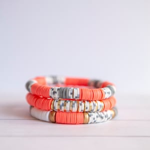 May include: Three stacked bracelets with coral, white, and gray beads. The bracelets have a geometric pattern and gold accents.