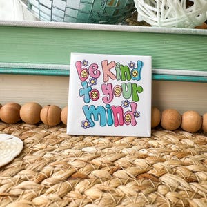 May include: A square white tile with the words "be Kind to your Mind" in colorful bubble letters and flower accents. The tile is on a woven mat with wooden beads and decorative items in the background.