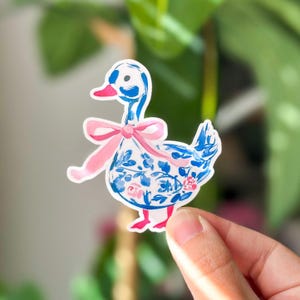 May include: A whimsical sticker featuring a duck with a blue floral pattern and a pink bow. The duck has a pink beak and feet. The sticker is held up by a hand in front of a blurred green background.