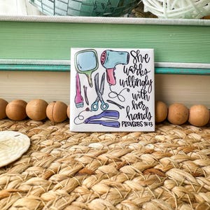 May include: A square ceramic tile featuring a hand-drawn illustration of hair styling tools in blue, pink, and purple. The tile includes a mirror, scissors, a hair dryer, and the text "She works willingly with her hands PROVERBS 31:13."