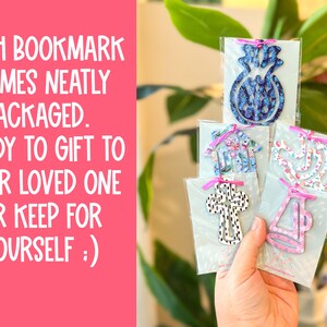 May include: Five different patterned bookmarks are displayed in their individual packaging. Each bookmark is made of wood and features a different design, including a pineapple, a house, a bird, a cross, and a megaphone. The bookmarks are ready to be gifted or kept for oneself.