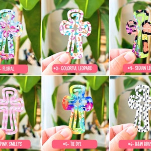 May include: Six different cross-shaped hair clips with various patterns, including floral, leopard print, tie-dye, and smiley faces. The clips are all made of plastic and have a white background.