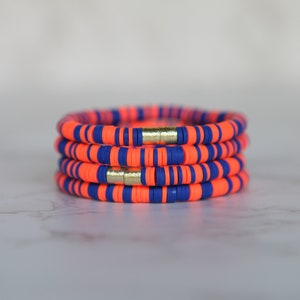 May include: Stack of four orange and blue beaded bracelets. Each bracelet features alternating orange and blue flat beads. The bracelets are held together with gold-colored magnetic clasps. The bracelets are stacked on a white marble surface.