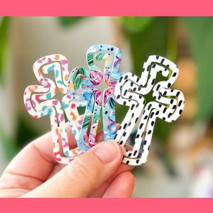 May include: Three metal cross-shaped hair clips with different patterned designs. One clip has a black and white polka dot pattern, another has a floral pattern with pink, blue, and green colors, and the third has a pink and white leopard print pattern.