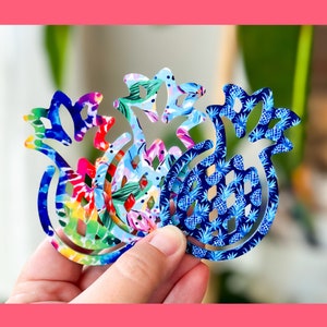 May include: Three pineapple-shaped cutouts with different colorful patterns. The cutouts are made of a thin, flexible material and are held in a hand.