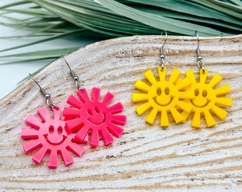 May include: A pair of yellow and pink smiley face sun earrings. The earrings are made of plastic and have a silver hook.