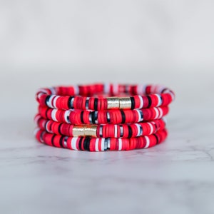 May include: A stack of four red, white, and black beaded bracelets with gold accents. The bracelets are made of small, round beads and are stacked on top of each other.