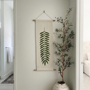 May include: A vertical off-white fabric wall hanging featuring a dark green fern leaf print. The textile is suspended from a light wood rod with a white cord and has a matching wood rod at the bottom. The piece is displayed on a plain wall.