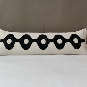 May include: A long white lumbar pillow with a black geometric pattern of circles connected by lines. The pillow is on a white bed.
