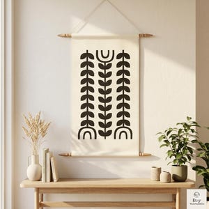 Könnte beinhalten: Ein rechteckiger Wandbehang aus cremefarbenem Canvas mit einem schwarzen Grafikdruck von drei vertikalen botanischen Stielen mit symmetrischen Blättern und bogenförmigen Basiselementen, aufgehängt an einer Holzstange mit einer Schnur.