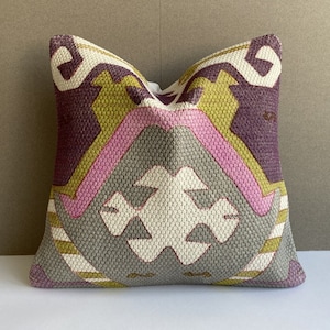 Könnte beinhalten: Ein dekoratives Kissen mit einem geometrischen Muster in den Farben Lila, Rosa, Grün und Grau. Das Kissen hat eine gewebte Textur und einen weißen Hintergrund.