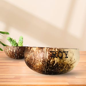 May include: Two brown coconut bowls, one with the name "ROY" engraved on the side. The bowls are on a wooden surface.