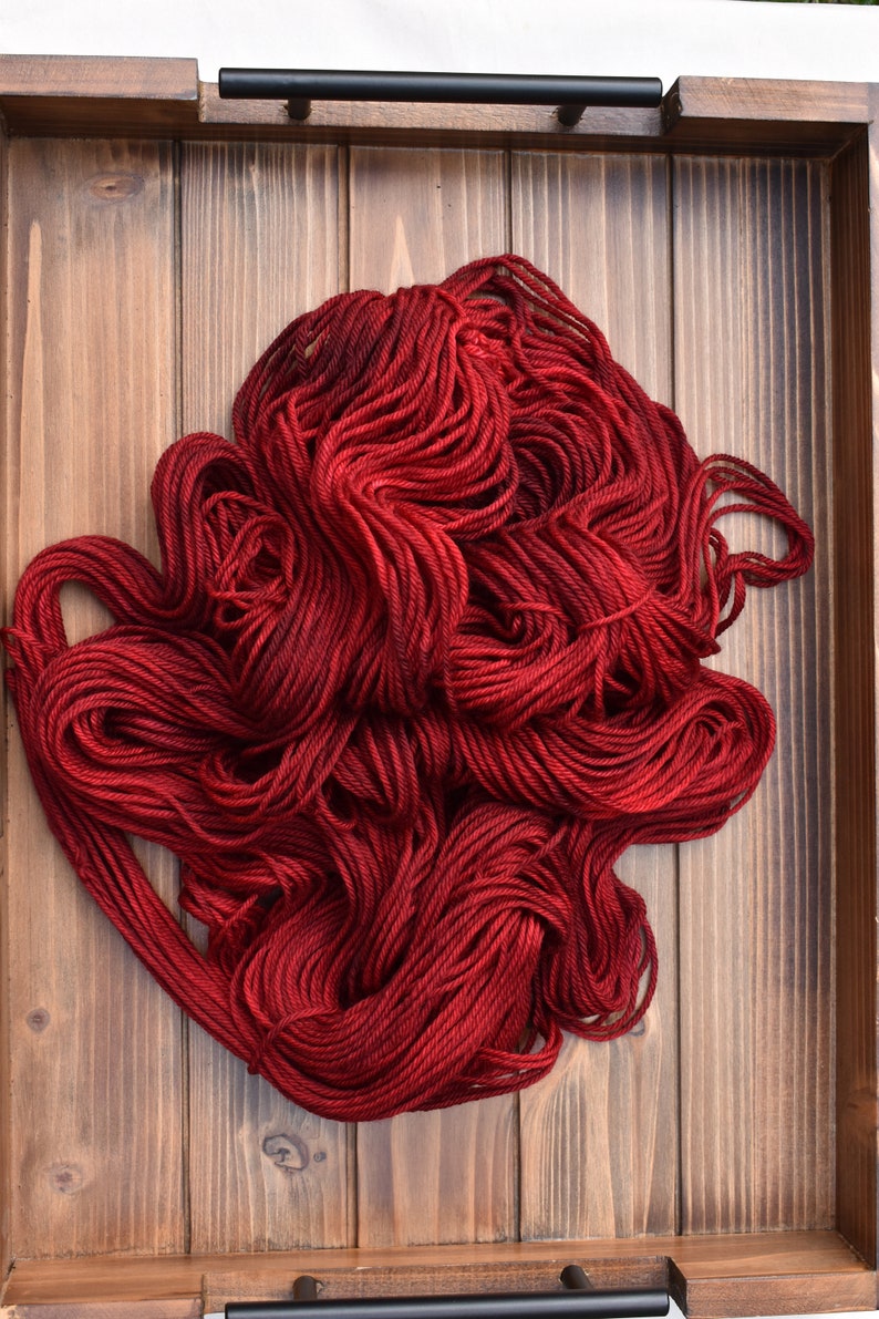 Puede incluir: Una madeja grande y suelta de hilo rojo oscuro con hebras retorcidas visibles. El hilo est&aacute; enrollado en un mont&oacute;n, mostrando varios tonos de carmes&iacute; y rojo oscuro a trav&eacute;s de las fibras, descansando dentro de una bandeja de madera rectangular con asas negras.