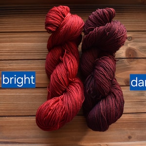 Puede incluir: Dos madejas de hilo mostradas una al lado de la otra. La madeja de la izquierda es de un color rojo brillante con la palabra "bright" impresa en un recuadro azul. La madeja de la derecha es de un rojo burdeos oscuro con la palabra "dark" en azul.