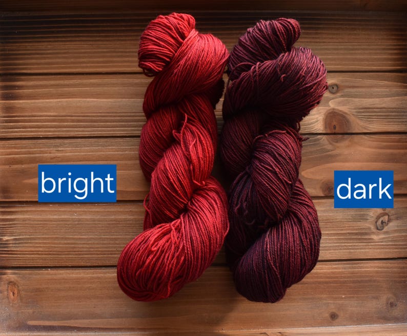 Puede incluir: Dos madejas de hilo mostradas una al lado de la otra. La madeja de la izquierda es de un color rojo brillante con la palabra "bright" impresa en un recuadro azul. La madeja de la derecha es de un rojo burdeos oscuro con la palabra "dark" en azul.