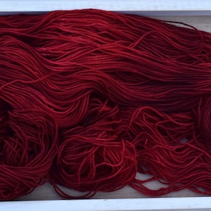 Puede incluir: Una madeja grande y suelta de hilo rojo carmes&iacute; profundo con una textura suave y ligeramente lustrosa. El hilo se compone de muchas hebras finas y continuas agrupadas en un arreglo org&aacute;nico y enredado, mostrando un color rico.
