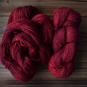 Puede incluir: Dos madejas de hilo de color rojo burdeos profundo con una textura suave y retorcida. El hilo se presenta en una disposici&oacute;n suelta y enrollada, mostrando el color rico y saturado y la estructura fina de las hebras de fibra.