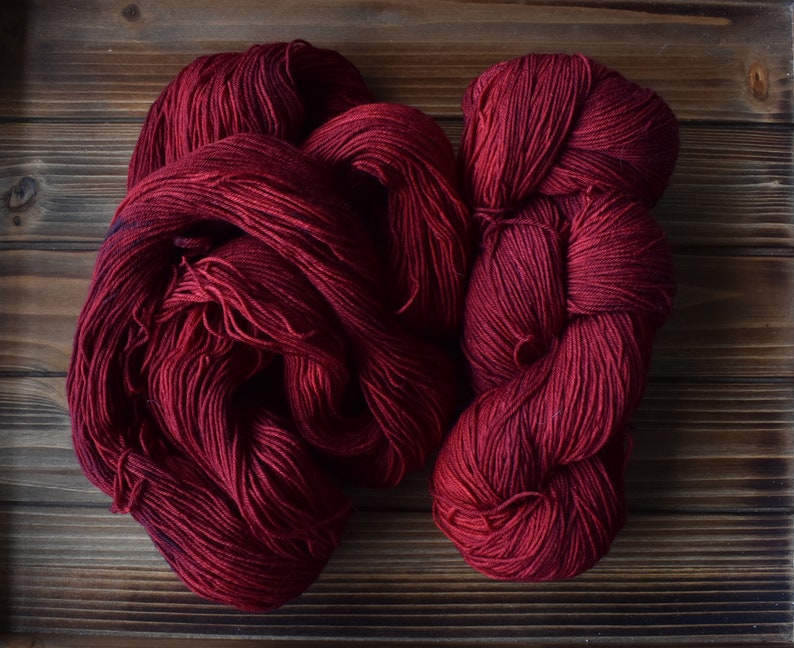 Puede incluir: Dos madejas de hilo de color rojo burdeos profundo con una textura suave y retorcida. El hilo se presenta en una disposici&oacute;n suelta y enrollada, mostrando el color rico y saturado y la estructura fina de las hebras de fibra.