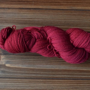 Puede incluir: Una madeja de hilo color burdeos intenso, retorcida en un bucle largo y continuo. El hilo presenta una textura consistente y finamente hilada con un acabado mate, mostrando un rico color rojo oscuro a lo largo de toda la fibra.
