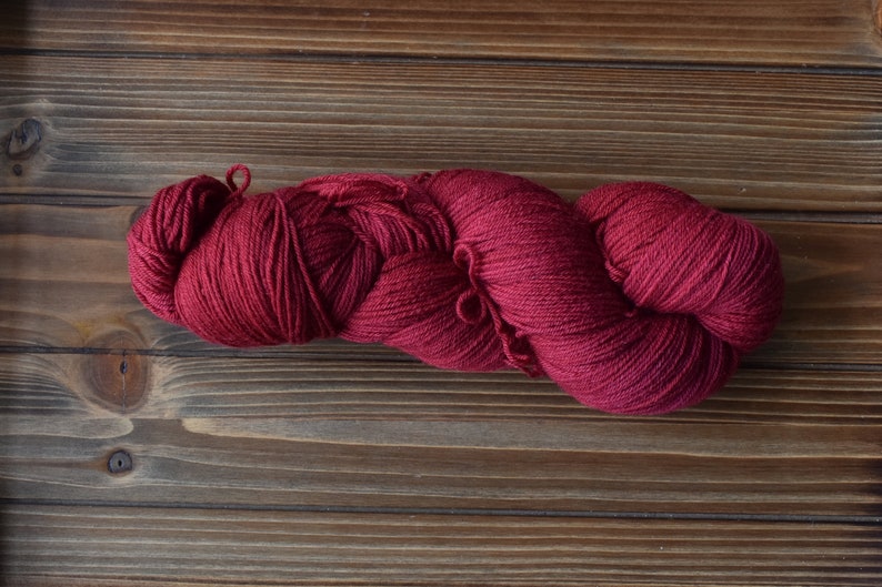 Puede incluir: Una madeja de hilo color burdeos intenso, retorcida en un bucle largo y continuo. El hilo presenta una textura consistente y finamente hilada con un acabado mate, mostrando un rico color rojo oscuro a lo largo de toda la fibra.