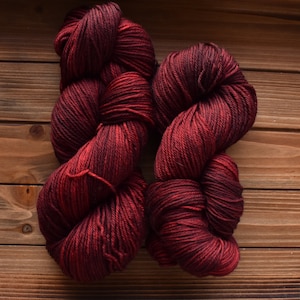 Puede incluir: Dos madejas de hilo con una paleta de colores variados y profundos de burdeos oscuro, carmes&iacute; y negro. La fibra est&aacute; retorcida en una estructura gruesa de varias hebras con un brillo visible, mostrando una rica mezcla de tonos rojos.