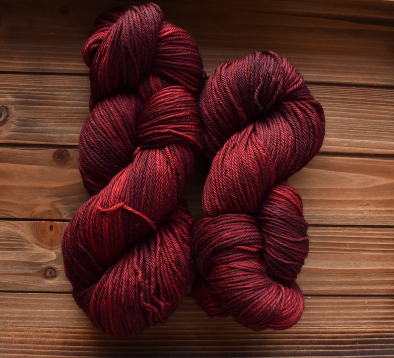 Puede incluir: Dos madejas de hilo con una paleta de colores variados y profundos de burdeos oscuro, carmes&iacute; y negro. La fibra est&aacute; retorcida en una estructura gruesa de varias hebras con un brillo visible, mostrando una rica mezcla de tonos rojos.