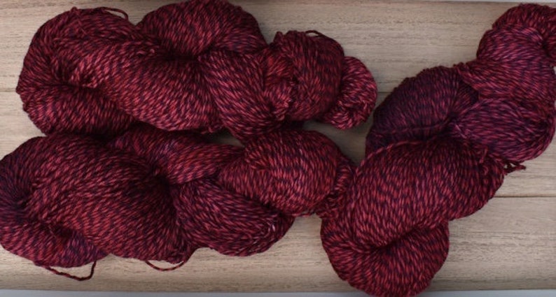 Puede incluir: Dos madejas de hilo con una construcci&oacute;n trenzada de m&uacute;ltiples cabos. El hilo presenta un patr&oacute;n de color jaspeado, mezclando fibras de color burdeos profundo y negro oscuro a lo largo de toda la longitud de los cabos.