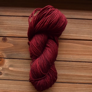 Puede incluir: Una madeja de hilo color burdeos intenso, retorcida firmemente en forma de ocho. El hilo presenta una construcci&oacute;n lisa de m&uacute;ltiples cabos con un color rojo oscuro consistente en toda la longitud del haz de fibras.