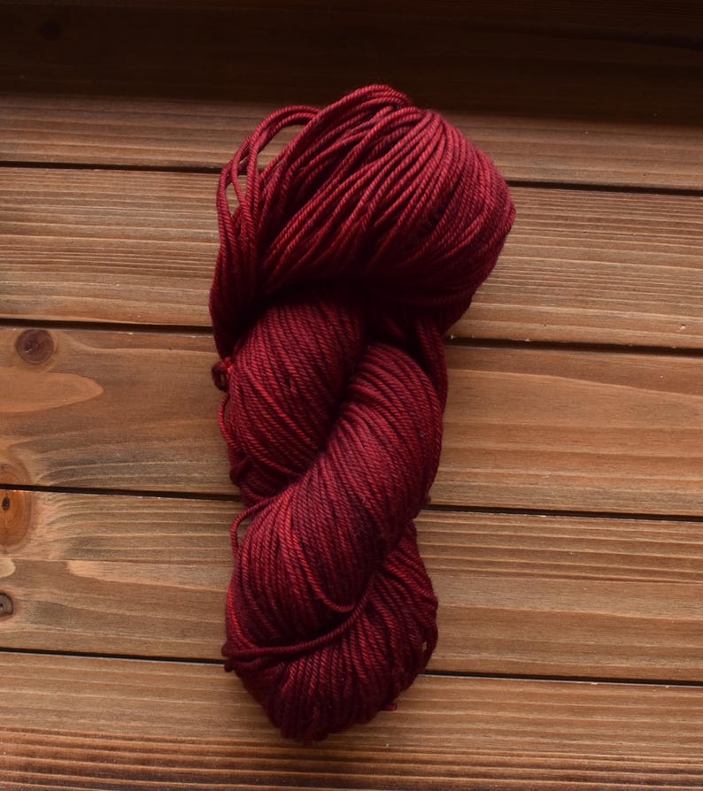 Puede incluir: Una madeja de hilo color burdeos intenso, retorcida firmemente en forma de ocho. El hilo presenta una construcci&oacute;n lisa de m&uacute;ltiples cabos con un color rojo oscuro consistente en toda la longitud del haz de fibras.