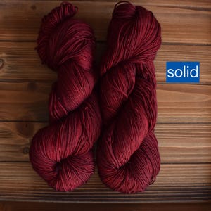 Puede incluir: Dos madejas de hilo color burdeos profundo con un acabado de color s&oacute;lido. El hilo est&aacute; trenzado en un paquete suave y texturizado, mostrando un tono rojo oscuro consistente en todas las fibras. Un gr&aacute;fico azul con el texto blanco solid.