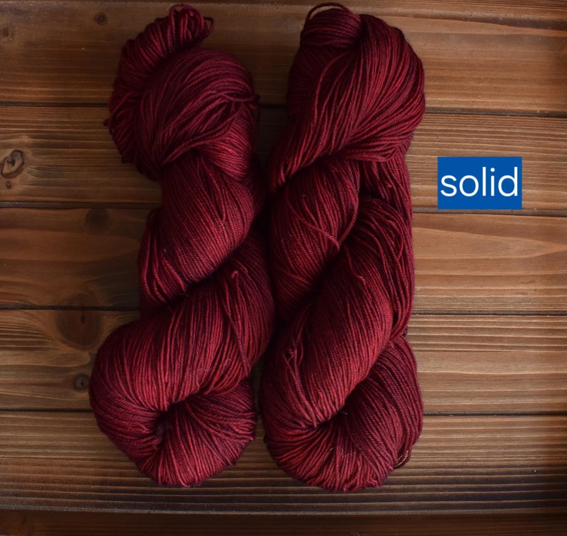 Puede incluir: Dos madejas de hilo color burdeos profundo con un acabado de color s&oacute;lido. El hilo est&aacute; trenzado en un paquete suave y texturizado, mostrando un tono rojo oscuro consistente en todas las fibras. Un gr&aacute;fico azul con el texto blanco solid.