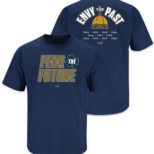May include: Navy blue t-shirt with gold text that reads "Fear the Future" and a green shamrock. The back of the shirt has gold text that reads "Envy the Past" with a gold dome and years listed.