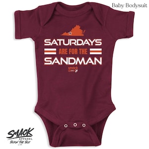 May include: Burgundy baby bodysuit with a white graphic that says "Saturdays Are For The Sandman" with a small orange Virginia state outline above the text. The graphic also includes two orange stripes. The text "smack apparel jr" is in small white letters below the graphic.